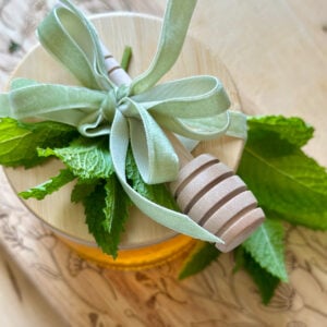 Mint honey in an embossed glass jar with a bamboo lid. The lid is covered with a light green velvet bow, a honey wand and sprigs of fresh spearmint.