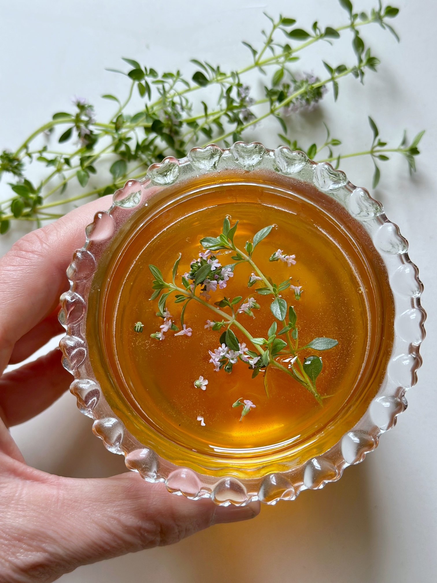 Hand holding a glass honey with thyme leaves and flowers floating.